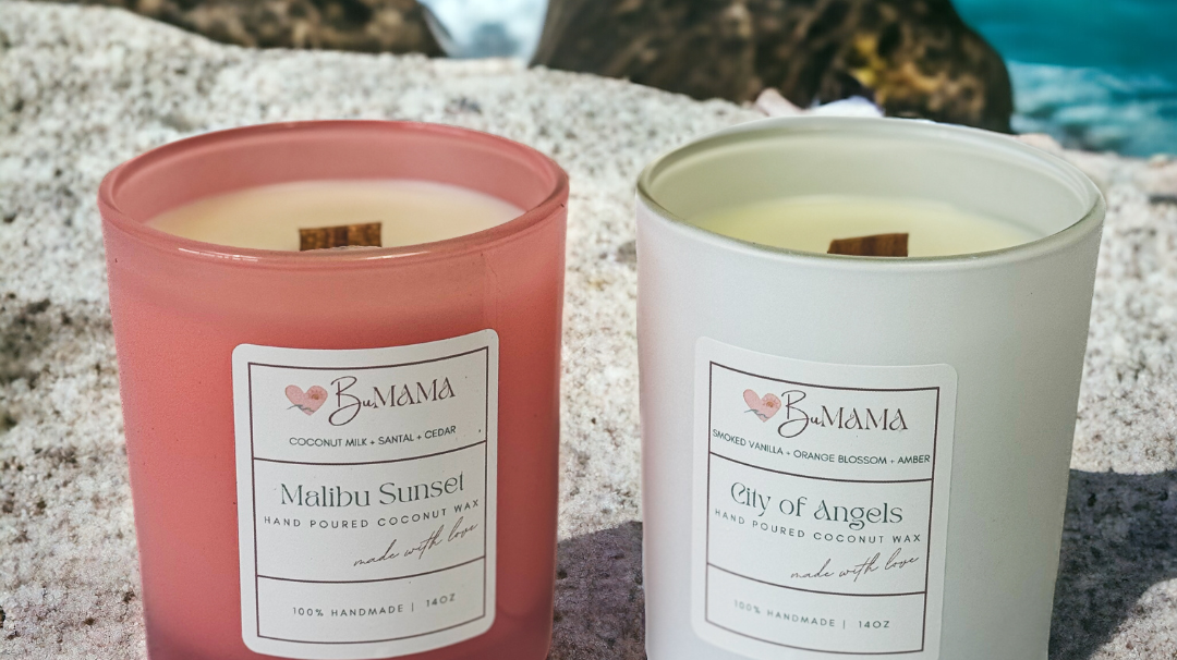 two candles one in a pink jar and one in a white jar sitting in the sand with water in the background